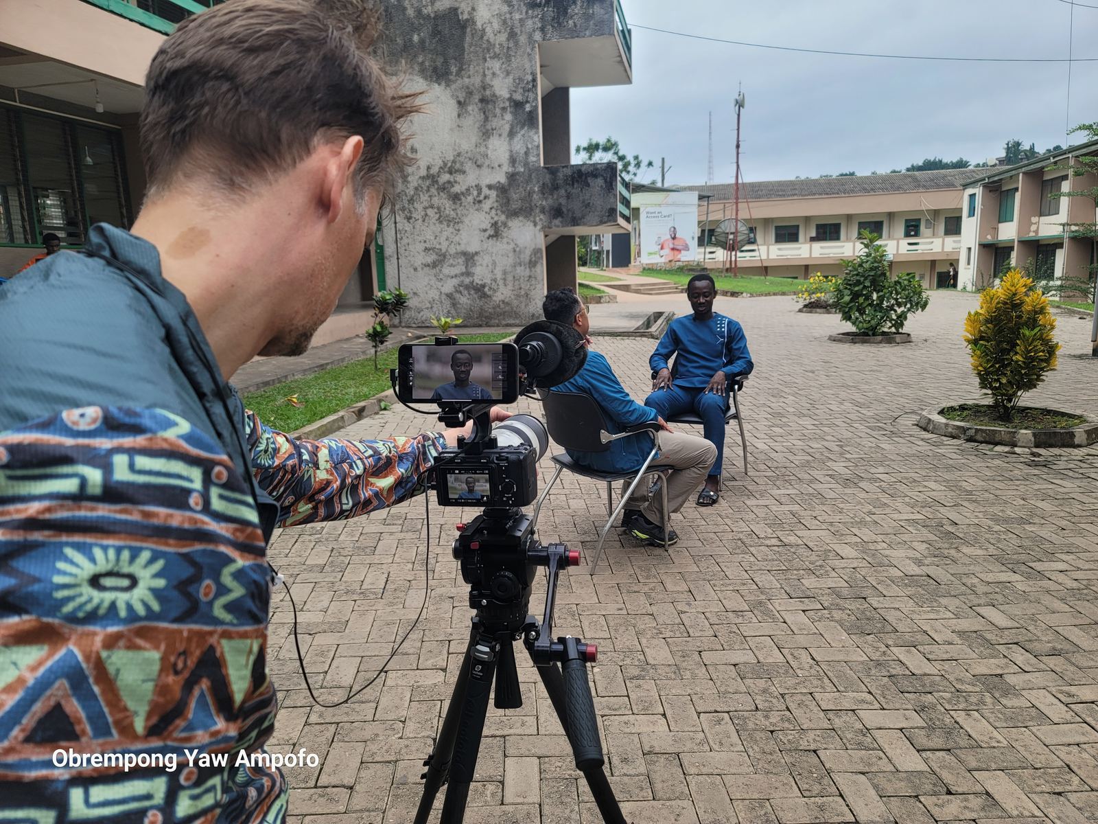 Crew filming a seated interviewee in a courtyard in Kenyasi